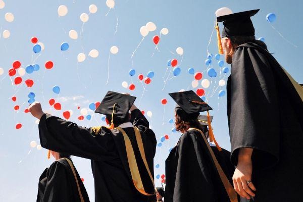 Graduates in black gowns and caps celebrate with balloons against a blue sky.