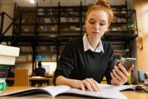 A young woman with red hair studies at a desk, looking at her phone and a book.