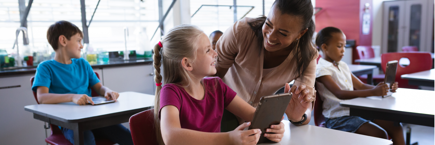 A teacher helps a student with a tablet in a classroom setting.