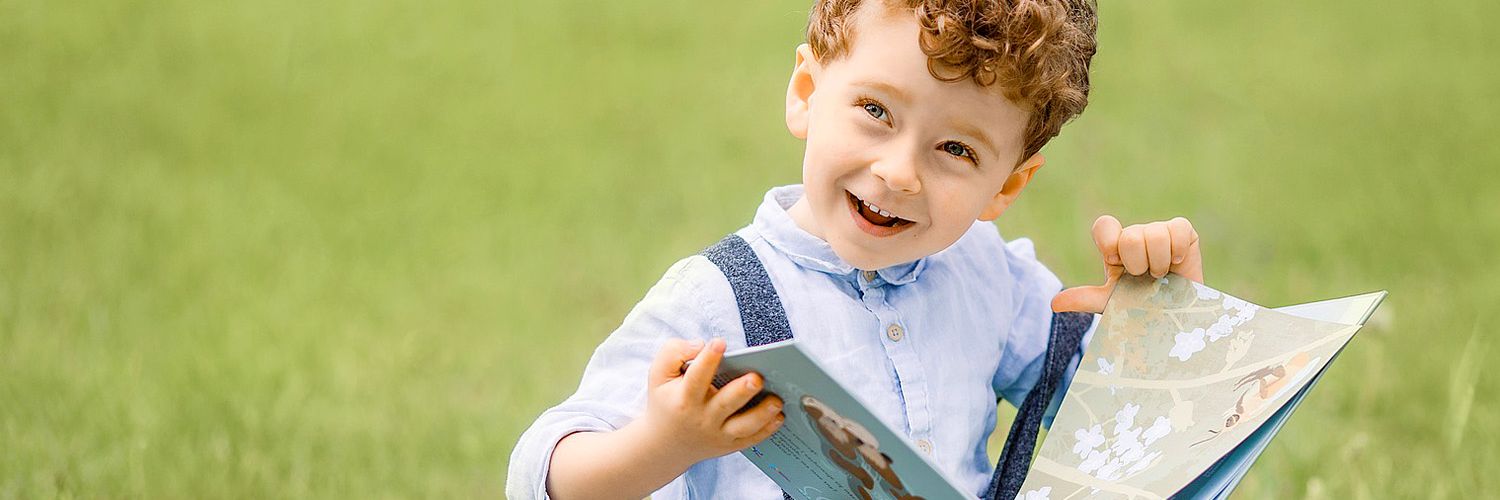 A young boy with curly red hair smiles brightly as he holds a book outdoors.