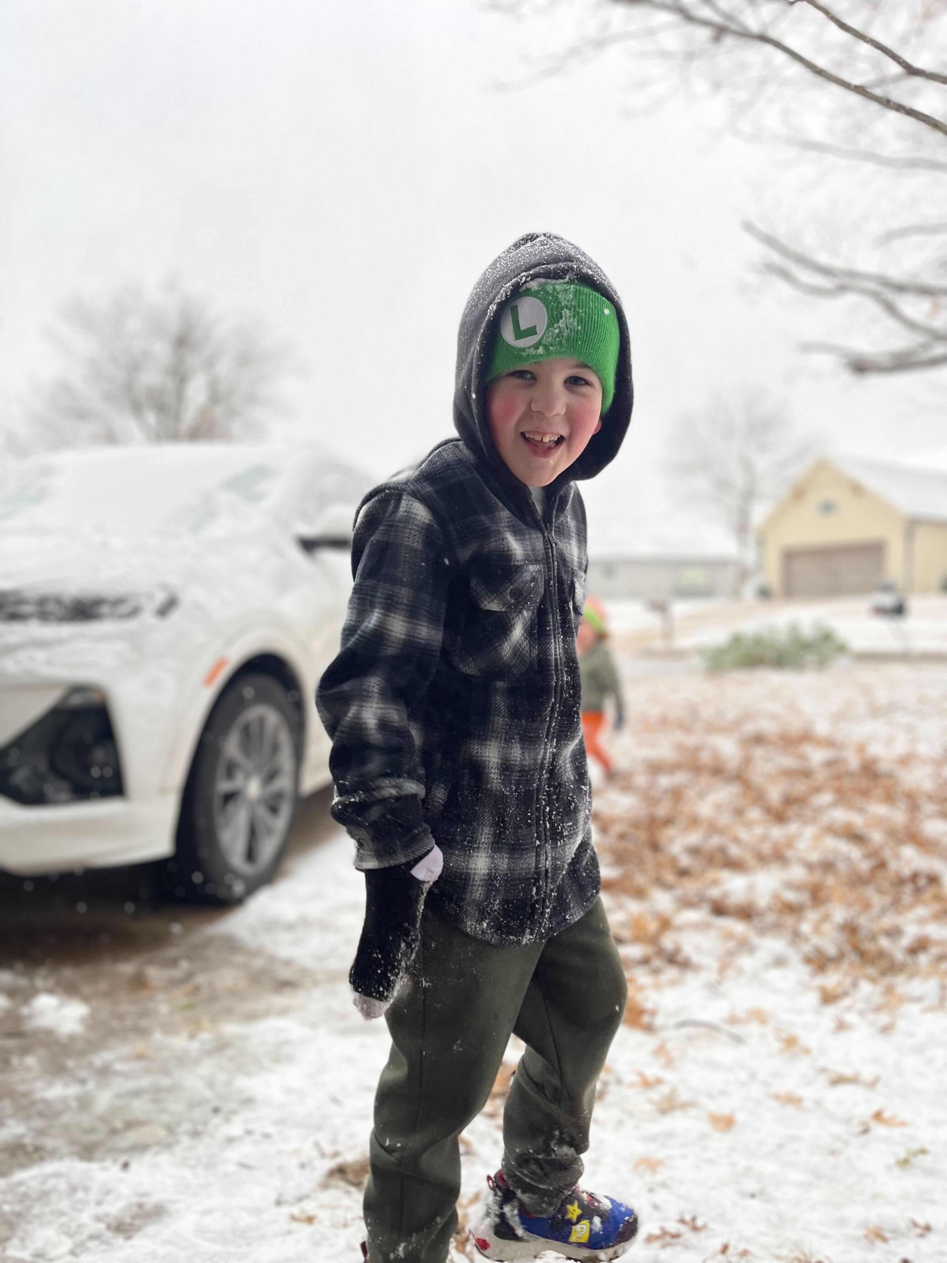A young boy wearing a green hat with an &amp;amp;#039;L&amp;amp;#039; on it smiles for the camera in a snowy driveway.