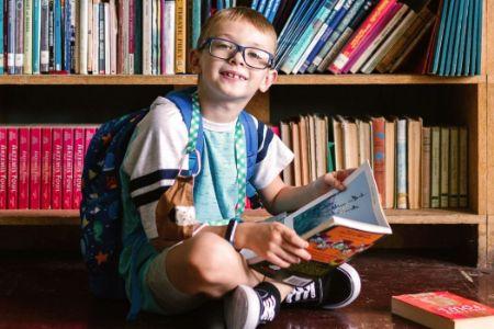 A young boy with glasses smiles while reading a book in a library.
