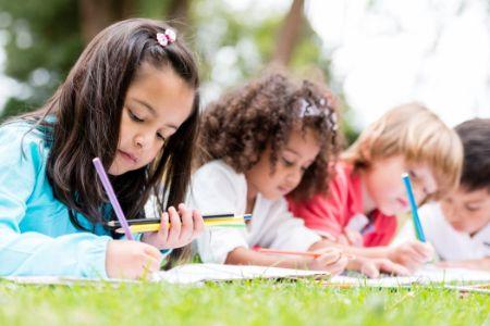 Children of diverse backgrounds concentrate on drawing or writing on paper outdoors.