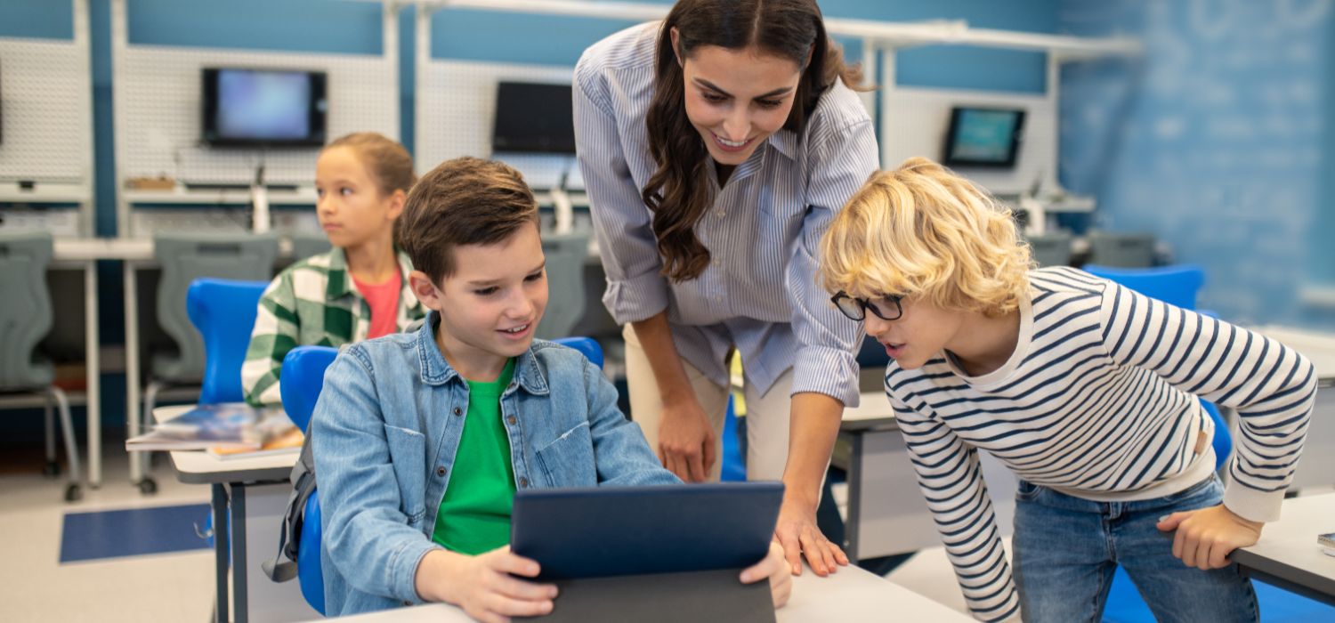 A teacher assists children with a tablet in a classroom setting.