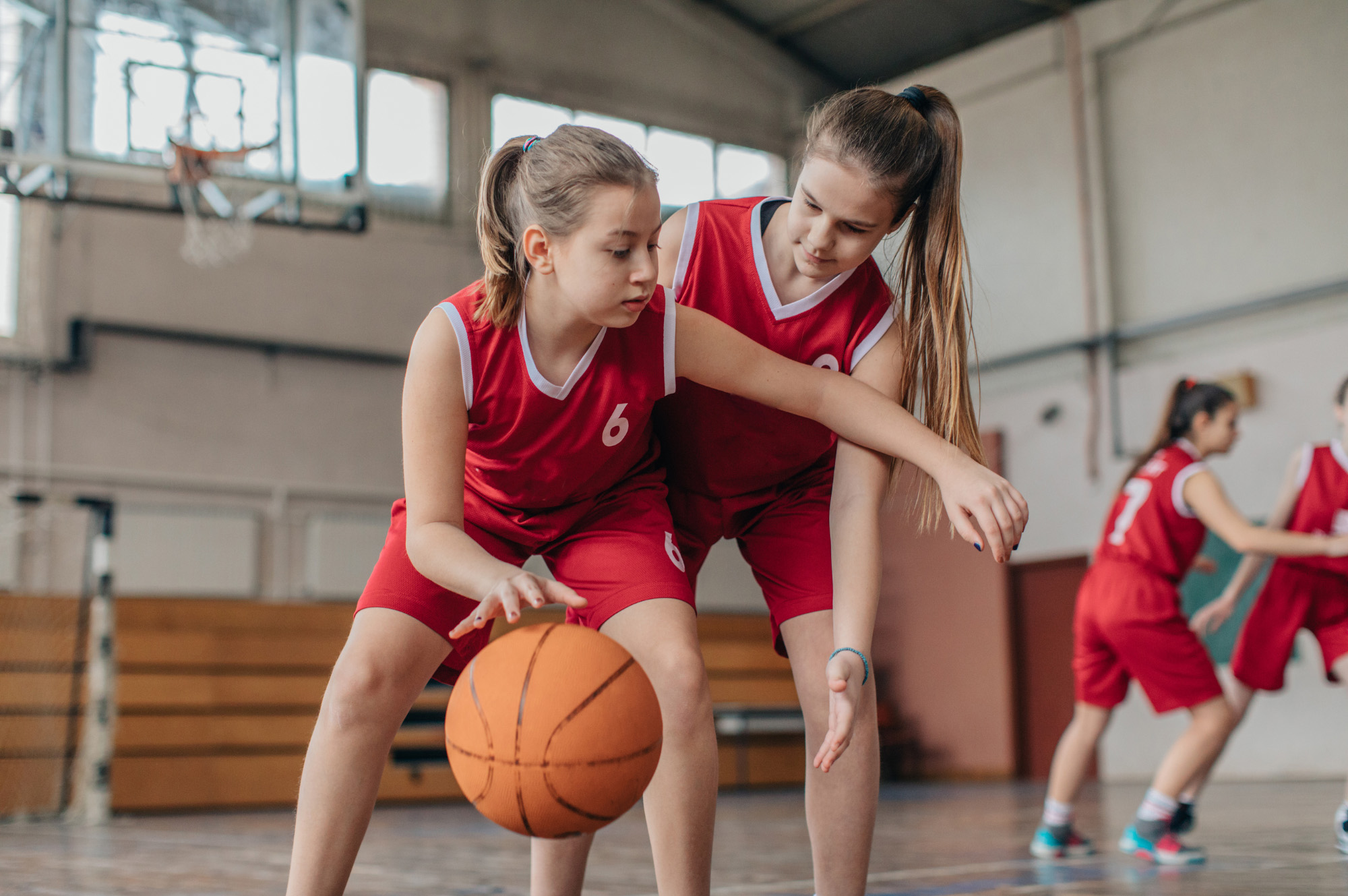 Two young girls in red basketball uniforms practice dribbling a basketball.